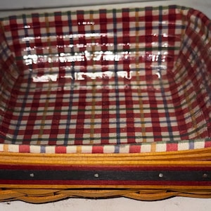 May include: A rectangular woven basket with a red, white, and blue plaid fabric lining. The basket has a yellow and black trim with small silver-colored studs. The basket is empty.