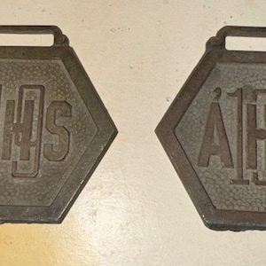 May include: Two hexagonal metal medallions with a watch fob attachment. Both medallions feature the text "AHS" with the number "1" in the center. The first medallion has the number "1" above the "A" and the second medallion has the number "2" above the "A".