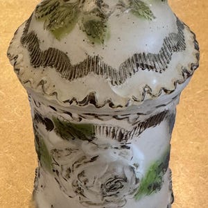 May include: White ceramic jar with a lid, decorated with green and brown floral designs. The jar has a scalloped edge and a raised floral design on the side.
