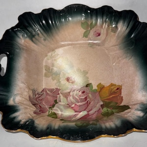 May include: An antique ceramic serving dish with a scalloped edge and two handles. The dish features a floral design with pink, yellow, and white roses on a cream-colored background, framed by a dark green border with gold trim.