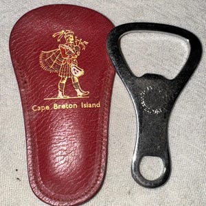 May include: A red leather bottle opener case with a gold image of a man in a kilt playing bagpipes. The case is labeled "Cape Breton Island". The bottle opener is silver and has a circular design on the back.