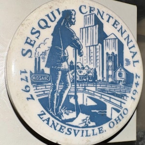 May include: A blue and white ceramic commemorative plate with the text "Sesqui Centennial 1797 Zanesville, Ohio 1947" and a depiction of a man standing with a rifle, a cityscape, and a bridge.