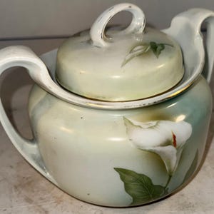 May include: A white porcelain sugar bowl with a lid, featuring a delicate floral design of white calla lilies and green leaves. The bowl has a gold trim around the rim and a slightly iridescent glaze.