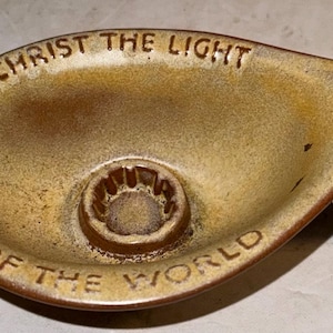 May include: A brown ceramic dish with a handle. The dish has a small hole in the center and the words "Christ the Light of the World" are inscribed around the rim.