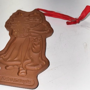 May include: A brown, Christmas-themed ornament in the shape of Santa Claus, with a red ribbon for hanging. The ornament has the words "Kris Kringle" at the bottom. The Santa Claus figure is wearing a long coat and holding a bag.