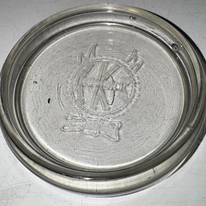 May include: A clear glass jar lid with a circular shape. The lid has a raised rim and a central embossed design featuring the letters "K" and "C" within a decorative emblem. The glass is transparent.