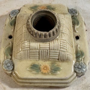 May include: A square, cream-colored ceramic ceiling fixture with a woven pattern and floral accents. The fixture has a metal socket for a light bulb.