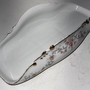 May include: A white porcelain dish with a curved, scoop-like shape. The rim is trimmed with gold and features a floral pattern with pink roses and green leaves. The dish has a scalloped edge.