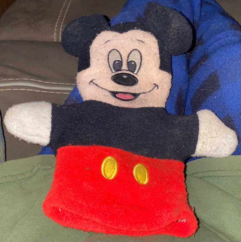 May include: A red, black and white plush Mickey Mouse hand puppet. The puppet has a black nose, two yellow buttons and a big smile.