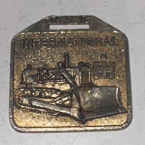 May include: A rectangular, gold-toned metal badge featuring the word "INTERNATIONAL" above a detailed relief of a bulldozer. The badge has a keyhole-shaped top with the letters "H S D" visible. The bulldozer is the central focus.