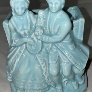 May include: A light blue ceramic planter shaped like a couple sitting on a bench. The woman is wearing a long dress and the man is wearing a long coat. The planter has a rectangular opening in the center for planting.