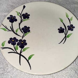May include: A round, off-white plate with a floral design. The plate features dark purple flowers with yellow centers and green leaves, arranged on black stems. The flowers are painted on the left and right sides of the plate.