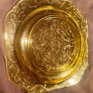 May include: A square, yellow-tinted glass plate with an ornate, embossed floral design. The plate has a scalloped edge and a central circular pattern. The glass has a slight iridescence, reflecting light.