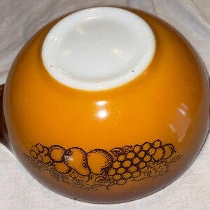 May include: An amber-colored Pyrex bowl with a white interior and brown handles. The exterior features a brown fruit design. This vintage bowl is ideal for serving or food preparation.