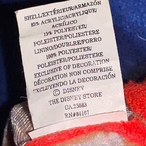 May include: A white tag with black text detailing the fabric composition of a garment. The tag reads "SHELL/EXT&Eacute;RIEUR/ARMAZ&Oacute;N 85% ACRYLIC/ACRYLIQUE/ACR&Iacute;LICO 15% POLYESTER/POLEISTER/POLIESTERE LINING/DOUBLRE/FORRO 100% POLYESTER/POLEISTER/POLIESTERE EXCLUSIVE OF DECORATION D&Eacute;CORATION NON COMPRISE EXCLUYENDO LA DECORACI&Oacute;N &copy; DISNEY THE DISNEY STORE CA.25083 RN#84167"