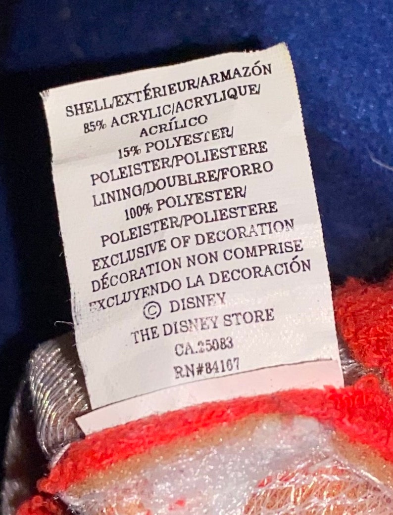 May include: A white tag with black text detailing the fabric composition of a garment. The tag reads "SHELL/EXT&Eacute;RIEUR/ARMAZ&Oacute;N 85% ACRYLIC/ACRYLIQUE/ACR&Iacute;LICO 15% POLYESTER/POLEISTER/POLIESTERE LINING/DOUBLRE/FORRO 100% POLYESTER/POLEISTER/POLIESTERE EXCLUSIVE OF DECORATION D&Eacute;CORATION NON COMPRISE EXCLUYENDO LA DECORACI&Oacute;N &copy; DISNEY THE DISNEY STORE CA.25083 RN#84167"