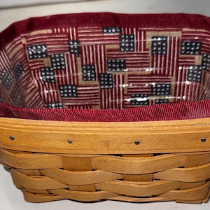 May include: Woven rectangular basket with a red fabric lining featuring a pattern of American flags. The basket has two brown leather handles and is made of light brown woven material.