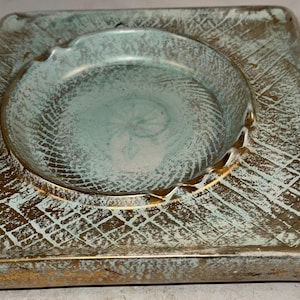 May include: Square ceramic ashtray with a green and gold glaze. The ashtray has a deep, round well in the center.