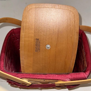 May include: A red and wood Longaberger basket with a wood lid. The lid has the text "Longaberger WoodCrafts  777  221-2211" printed on it.