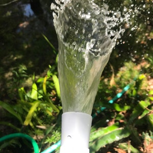 Puede incluir: Primer plano de una boquilla de plástico blanca que rocía agua, creando un rocío de gotas de agua. La boquilla está conectada a una manguera de jardín.