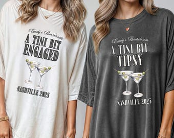 May include: Two oversized t-shirts, one cream and one charcoal, featuring the text "Emily's Bachelorette A Tini Bit Engaged Nashville 2025" and "Emily's Bachelorette A Tini Bit Tipsy Nashville 2025". Each shirt has a martini graphic.