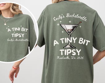 May include: Olive green oversized t-shirt with white text that reads "A TINI BIT TIPSY" and "Emily's Bachelorette". The back of the shirt features a martini glass graphic and the text "A TINY BIT TIPSY Nashville, In 2025".