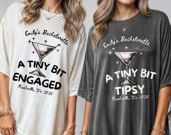 May include: Two oversized t-shirts, one cream and one charcoal gray, with the text "Emily's Bachelorette" and a cocktail glass graphic. The cream shirt says "A Tiny Bit Engaged" and the gray shirt says "A Tiny Bit Tipsy". Both shirts include "Nashville, TN 2025".
