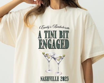 May include: Cream-colored t-shirt with the text "Emily's Bachelorette, A Tini Bit Engaged, Nashville 2025" in green and black. The shirt features a graphic of two martini glasses with olives. The shirt is a relaxed fit.