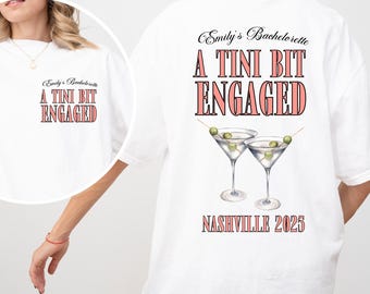 May include: White t-shirt with the text "Emily's Bachelorette" and "A TINI BIT ENGAGED" in a pink and black font. The back of the shirt features two martini glasses and the text "Nashville 2025".