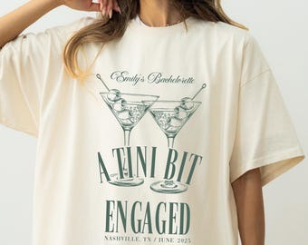 May include: A white t-shirt with a green graphic of two martini glasses with olives and the text "Emily's Bachelorette A Tini Bit Engaged Nashville TN / June 2023"