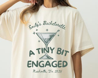 May include: Cream-colored t-shirt with the text "Emily's Bachelorette" above a martini glass graphic. Below the graphic, the text reads "A Tiny Bit Engaged" and "Nashville, Tn 2025". The shirt has short sleeves and a relaxed fit.