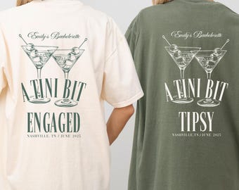 May include: Two oversized t-shirts, one cream and one olive green, with a martini graphic. The cream shirt says "A Tini Bit Engaged" and the green shirt says "A Tini Bit Tipsy". Both shirts have "Emily's Bachelorette" and the location and date printed on them.