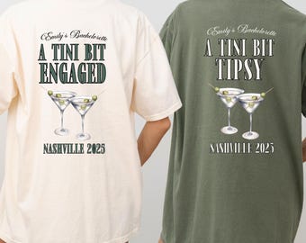 May include: Two t-shirts, one cream and one olive green, with text and martini glass graphics. The cream shirt says "Emily's Bachelorette, A Tini Bit Engaged, Nashville 2025." The green shirt says "Emily's Bachelorette, A Tini Bit Tipsy, Nashville 2025."