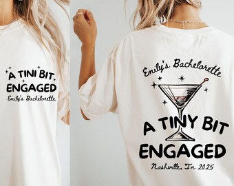 May include: Cream-colored t-shirt with black text that reads "A TINI BIT ENGAGED" and "Emily's Bachelorette" on the front. The back of the shirt features a martini glass graphic and the text "Emily's Bachelorette", "A TINY BIT ENGAGED", and "Nashville, In 2025".