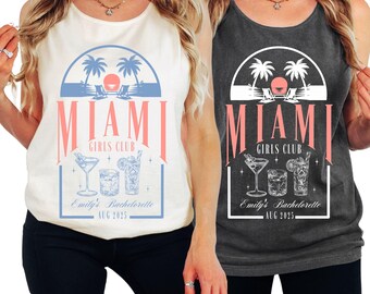 May include: Two tank tops, one cream and one charcoal, featuring a Miami Girls Club design. The design includes palm trees, cocktails, and the text "Emily's Bachelorette AUG 2025" in a rectangular frame. The shirts are sleeveless.