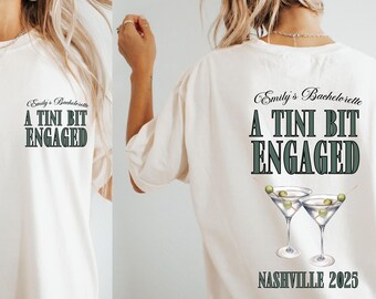 May include: Cream-colored t-shirt with the text "Emily's Bachelorette, A Tini Bit Engaged" in green and black. The back of the shirt features martini glasses and "Nashville 2025". A fun shirt for a bachelorette party.