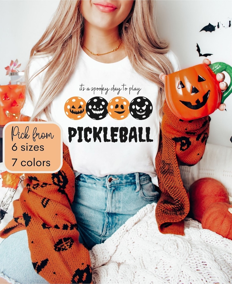 May include: White t-shirt with the text "it's a spooky day to play pickleball" and a row of four orange and black pickleball paddles with jack-o-lantern faces. The text "Pick from 6 sizes 7 colors" is in a white box.