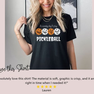 May include: Black t-shirt with a white graphic of three pickleball paddles with jack-o-lantern faces. The text "It's a spooky day to play PICKLEBALL" is on the shirt.