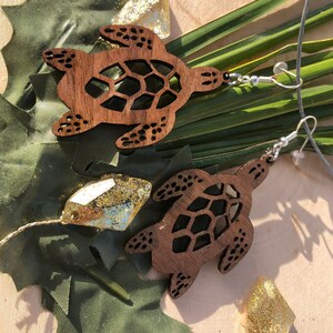 May include: A pair of wooden turtle earrings with a cutout design. The earrings are brown and have a natural wood finish. The turtles are facing the same direction and have a detailed design.