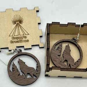 May include: A pair of wooden earrings featuring a wolf howling at the moon with trees in the background. The earrings are packaged in a small wooden box with the words "Ruppys Creations" on the lid.