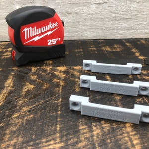 May include: Three grey plastic 3D printed brackets with the text "Deadmoose Tool Company" engraved on each. A red and black Milwaukee 25 ft tape measure is in the background.