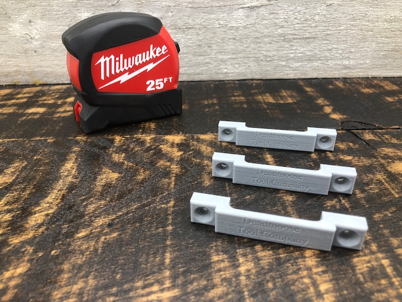May include: Three grey plastic 3D printed brackets with the text "Deadmoose Tool Company" engraved on each. A red and black Milwaukee 25 ft tape measure is in the background.