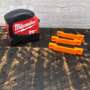May include: A red and black Milwaukee 25 ft tape measure and three orange plastic wall mounts.