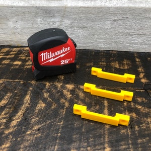 May include: A red and black Milwaukee 25 ft tape measure and three yellow plastic wall mounts.