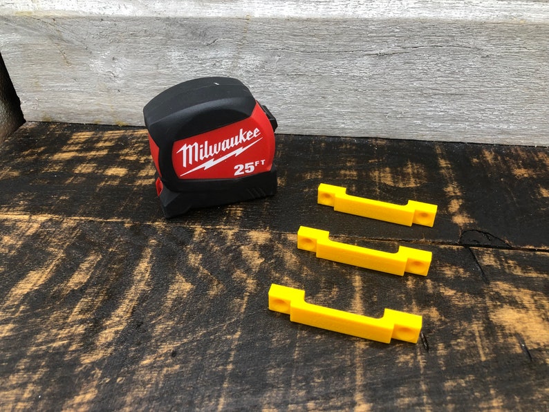 May include: A red and black Milwaukee 25 ft tape measure and three yellow plastic wall mounts.