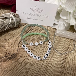 Può includere: Due braccialetti di perline e una collana a catena d'argento. Un braccialetto è verde chiaro, l'altro è verde più chiaro. La collana recita "DIVINE ENERGY" in perline di lettere bianche. Un biglietto da visita è sullo sfondo.