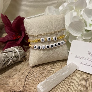 Op de afbeelding: Drie armbanden met kralen, gouden accenten en het woord "ABUNDANCE". Eén armband heeft de cijfers "888". De armbanden worden gepresenteerd op een beige kussen, met een witte saliebundel en een selenietstaaf.