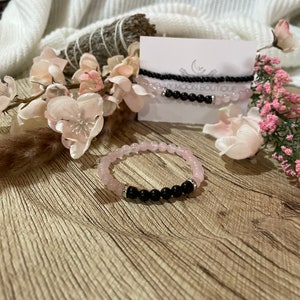 May include: Rose quartz and black onyx beaded bracelets. One bracelet features rose quartz beads, while another combines rose quartz and black onyx. A third bracelet is made of black onyx beads. The bracelets are displayed with pink flowers.