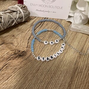 May include: Two beaded bracelets and a silver chain necklace. One bracelet features the word "STABILITY" spelled out in white letter beads. The other bracelet has the number "444". Both bracelets have light blue beads.