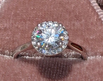 May include: A silver ring with a large, round, clear diamond surrounded by a halo of smaller diamonds. The ring is in a pink velvet ring box.